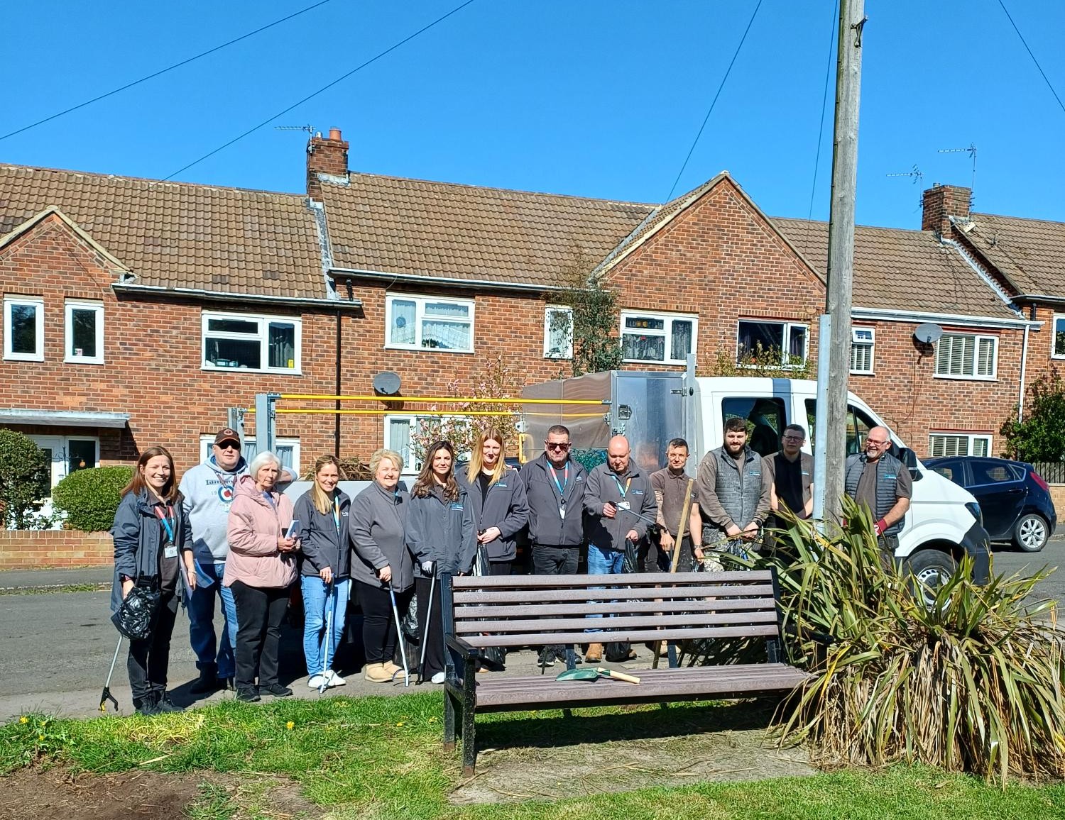 Thirteen people stood in front of a can and houses. A bench and plant in the foreground